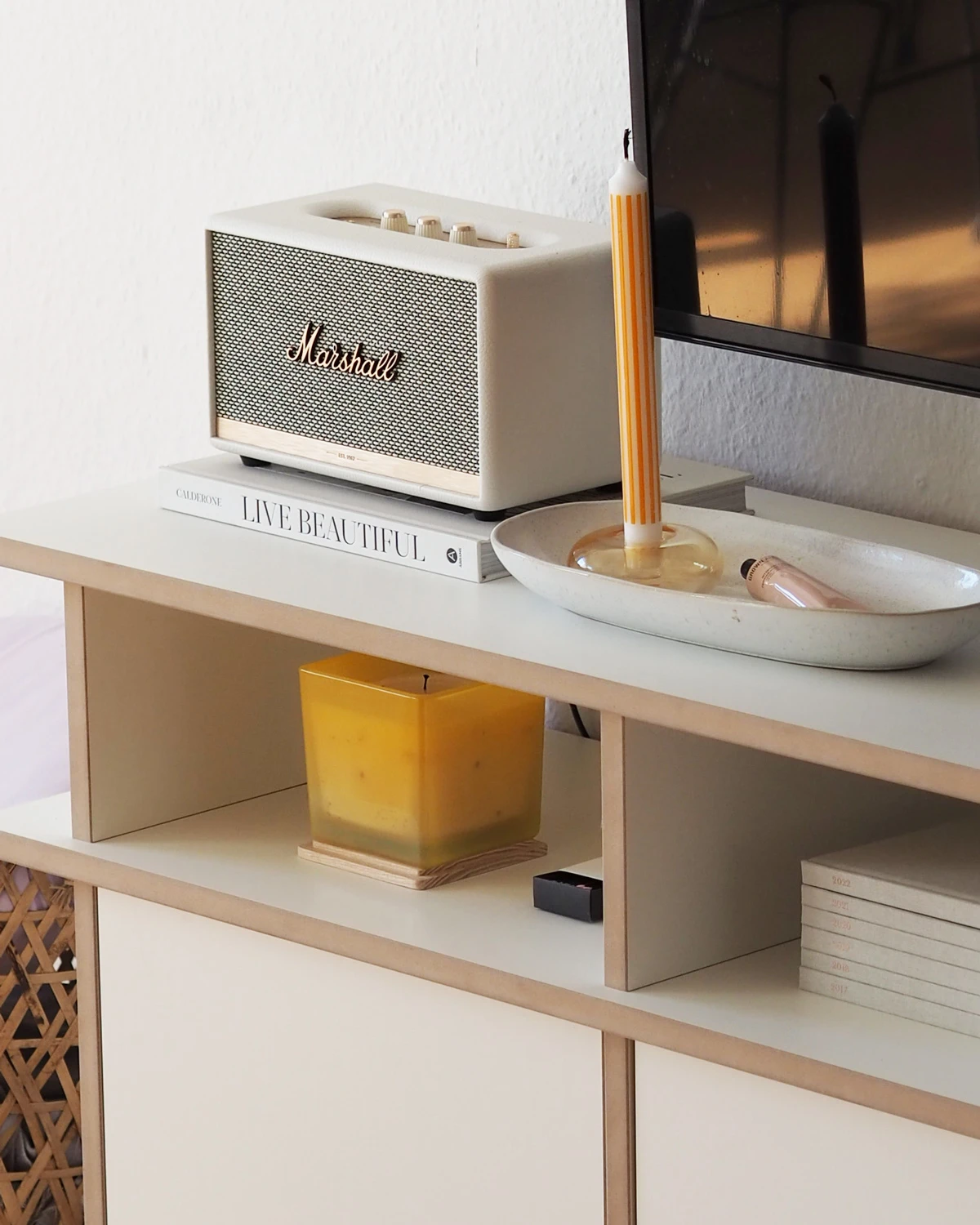 Gros plan sur un meuble TV bas blanc avec bordure en bois, des compartiments ouverts, une enceinte, une bougie et des objets décoratifs